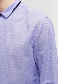 Jasnofioletowa koszula z wzorzystą teksturą, kołnierzykiem na guziki oraz granatowym akcentem na kołnierzu. Tkanina wydaje się gładka i lekko błyszcząca.