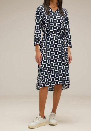 Granatowa sukienka z białym geometrycznym wzorem, w serek, z rękawami 3/4, z paskiem w talii, długości do kolan, w zestawie z białymi trampkami. Gładka tekstura.