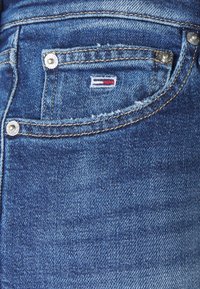 Vaqueros de mezclilla en un tono azul medio con una textura desgastada. Presentan un bolsillo delantero con un pequeño logo bordado y detalles de herrajes plateados.