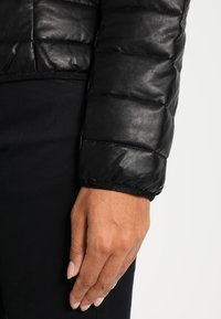 Svart pufferjacka med quiltad textur, tillverkad av läder. Manschetten är elastisk med en slät yta. Svart tyg under.