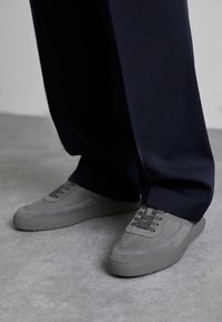 Gray suede sneakers with textured overlays and lace-up design. Rounded toe, flat soles, and visible stitching detail. Paired with dark trousers.