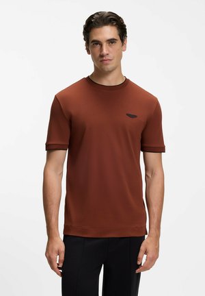 Giovane uomo con capelli scuri indossa una maglietta a maniche corte tinta unita di colore rosso mattone e pantaloni neri, in piedi davanti a uno sfondo bianco.