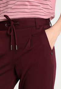 Burgundské kalhoty s elastickým pasem a stahovací šňůrkou, bočními kapsami a černým knoflíkem. Texturovaná tkanina s přiléhavým střihem.