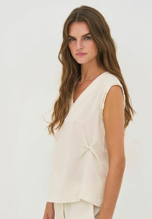 Hosszú, hullámos barna hajú nő, aki ujjatlan, krémszínű, V-nyakú felsőt visel, egyszerű világos háttér előtt áll.