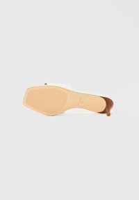 Sandala na potpeticu s bijelim gornjim dijelom od sintetičkog materijala, otvorenim prstima i tankom smeđom drvenom potpeticom. Teksturirana bež gumena potplat s linijama za bolje prianjanje.