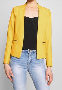 Blazer jaune au design sans col, poches avant zippées et texture lisse, associé à un haut noir et un jean bleu clair.