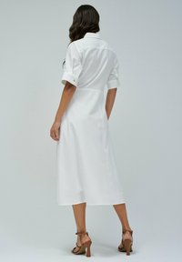 Abito camicia bianco con maniche corte arrotolate, di lunghezza media, realizzato in tessuto leggero. Il retro presenta una vestibilità sartoriale, che rivela una superficie liscia.