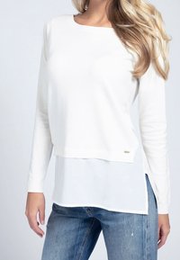 Maglione bianco a maniche lunghe con un aspetto a strati, caratterizzato da una consistenza morbida, scollatura rotonda e fessure laterali, abbinato a jeans azzurri chiari.