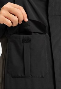 Hand die een open flap van een zwart stoffen zak met klittenband sluitingen vasthoudt op een zwart buitenkledingstuk.