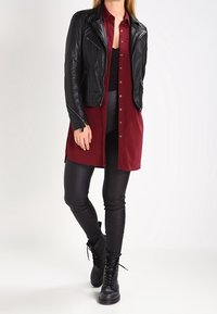 Donna che indossa una giacca di pelle nera sopra una camicia lunga buttons bordeaux, leggings neri e stivaletti neri con lacci, in piedi con le gambe incrociate.