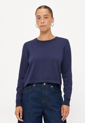 Navy blue cropped sweater with long sleeves, ribbed neckline and cuffs, made from a smooth knit fabric. Worn with high-waisted dark denim jeans.