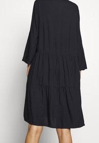 Svart, lager-på-lager klänning med trekvartsärmar. Har ett glättat tyg, lös passform och subtila rynkor vid midjan och nederkanten.