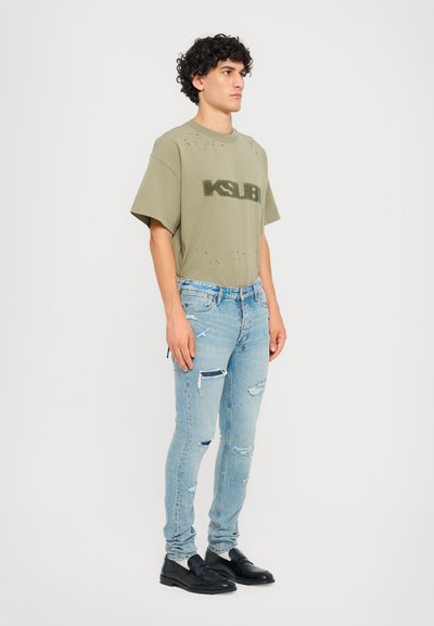 Ympäröivän oliivinvihreä ylisuurennettu t-paita, jossa on kuluneita yksityiskohtia ja "KSUBI"-logo, yhdistettynä vaaleansinisiin revittyihin skinny-farkkuihin ja mustiin loafer-kengiin.