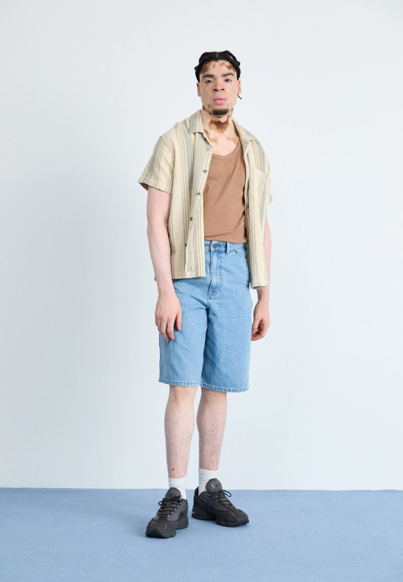 Chemise à manches courtes à rayures sur un débardeur marron, associée à un short en denim bleu clair et des baskets noires avec des chaussettes blanches.