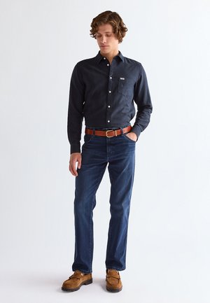 Junger Mann steht und trägt ein dunkles langärmliges Hemd mit Knöpfen, blaue Jeans, braunen Gürtel und braune Wildleder-Loafer vor einem einfarbigen Hintergrund.