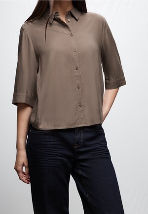 Vrouw die een bruine blouse met knopen en halflange mouwen en donkerblauwe jeans draagt, staand met één hand in de zak tegen een effen achtergrond.