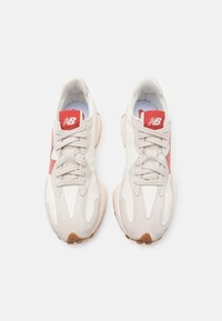 Ein Paar weiße und beige New Balance Sneakers mit roten Logopatches und dicken beigen Sohlen, von oben auf einem weißen Hintergrund betrachtet.