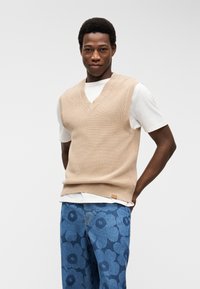 Άνδρας που φοράει μπεζ πλεκτό γιλέκο πάνω από λευκό μπλουζάκι και μπλε τζιν με μεγάλα σκουρόχρωμα λουλουδένια μοτίβα, στέκεται με τα χέρια πίσω από την πλάτη.