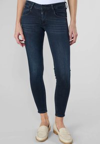 Jean skinny bleu foncé avec une taille mi-haute, en denim, avec une légère décoloration et un design classique à cinq poches, associés à des mocassins de couleur claire.