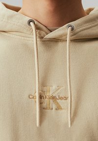 Sudadera con capucha beige hecha de tela suave, que presenta un gran logotipo bordado "CK" y cordones de color claro con puntas de metal.