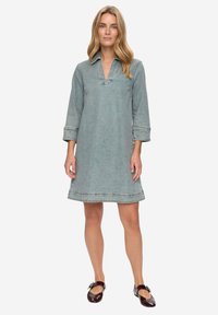Denimové šaty světle modré barvy s výstřihem do V, tříčtvrtečními rukávy a olemovaným spodním lemem. Ploché švy a minimální zdobení zdůrazňují jejich jednoduchost.