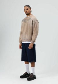 Beige sweatshirt met druppelende tekst, spijkershort en zwarte schoenen met roze veters. Model staat tegen een lichtgrijze achtergrond.