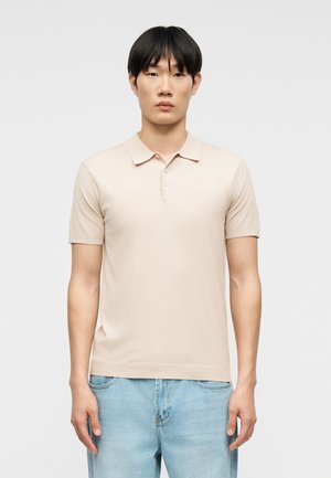 Junger Mann mit glatten schwarzen Haaren, der ein beige-farbenes kurzärmeliges Poloshirt und hellblaue Jeans trägt, vor einem einfarbigen Hintergrund stehend.