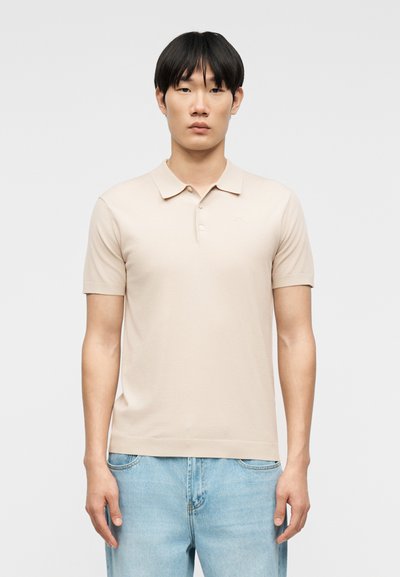 Giovane uomo con capelli lisci e neri che indossa una polo beige a maniche corte e jeans azzurri, in piedi contro uno sfondo semplice.