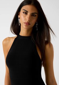 Abito nero a coste senza maniche con collo alto. La modella ha lunghi capelli scuri, indossa orecchini pendenti con cristalli e ha un incarnato liscio.