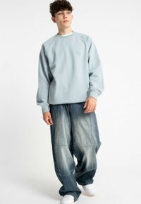 Jasnoniebieska bluza z okrągłym dekoltem i rękawami raglanowymi, w zestawieniu z luźnymi, wyblakłymi dżinsami z wieloma panelami. Białe sneakersy.