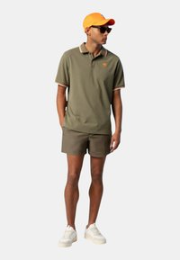 Polo verde oliva con colletto a strisce arancioni, abbinato a pantaloni corti marroni e scarpe bianche. Gli accessori includono un cappellino arancione e occhiali da sole.