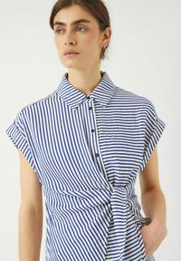 La camicia a righe presenta righe verticali blu e bianche, maniche corte, chiusura a bottoni, colletto e un dettaglio di vita legata.