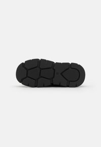 Black rubber shoe sole with a textured surface, featuring deep grooves and a jagged pattern for traction and stability.