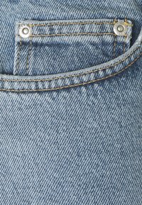 Gros plan sur un tissu en denim bleu montrant une petite poche à monnaie avec des rivets métalliques et des détails de couture jaunes.