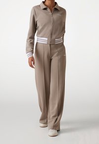 Beige jas met ritssluiting en bijpassende broek met witte strookaccenten op de manchetten en taille. Gemaakt van glad, rekbaar materiaal.