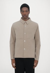 Tan jacket made of cotton blend fabric, featuring a collar, two front pockets, and silver snap buttons. The design is minimalist and structured.