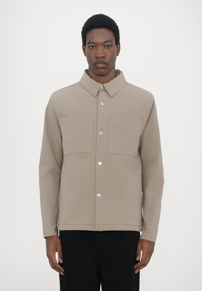 Tan jacket made of cotton blend fabric, featuring a collar, two front pockets, and silver snap buttons. The design is minimalist and structured.
