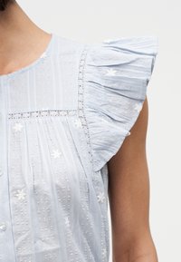 Blouse sans manches bleu clair avec épaules volantées, fleurs blanches brodées et bordure en dentelle portée sur le haut du torse d'une personne.