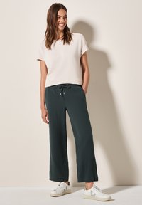 Light pink, short-sleeve top paired with dark green, loose-fitting, textured trousers. Worn with white sneakers featuring grey accents.