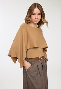 Poncho marrone lavorato a maglia con design a strati che cade su una maglietta marrone chiaro. Abbinato a pantaloni a vita alta a quadri e una cintura marrone.