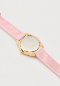 Goldfarbene Uhr mit rosa strukturiertem Silikonarmband, Edelstahlrückseite mit eingraviertem Guess-Logo und Angaben zur Wasserdichtigkeit.