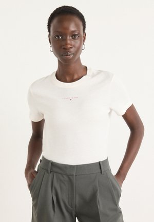 T-shirt en coton blanc avec un petit logo rouge "TOMMY JEANS" sur la poitrine, associé à un pantalon taille haute gris avec des plis.