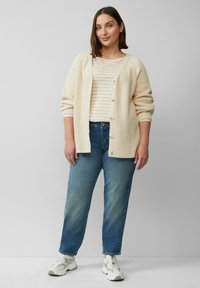 Lichtbeige gebreide cardigan met knopen, gedragen over een lichtgestreept shirt. Gecombineerd met medium-wash rechte jeans en witte sneakers.