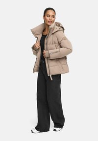 Beige pufferjack met een afneembare capuchon, een ritssluiting aan de voorkant en zijzakken, gecombineerd met zwarte wijde broek en witte sneakers.