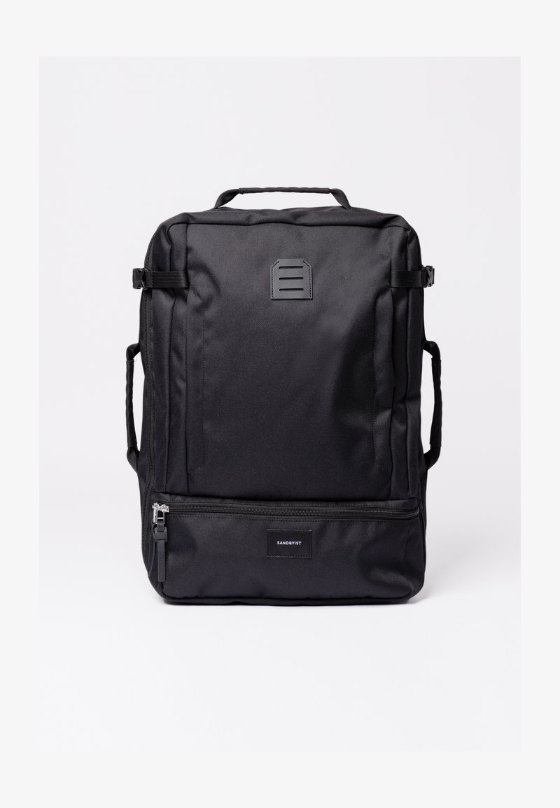Sac à dos rectangulaire noir avec poignée supérieure, sangles latérales, poche avant zippée et petit patch logo rectangulaire.