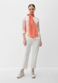 Gestreepte lange mouwen top in wit en koraal, gecombineerd met witte broeken en een gestructureerde koraal sjaal. Lichtgekleurde hoge sneakers maken de outfit af.