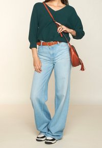 Suéter de canalé verde oscuro con escote en V, jeans de pierna ancha de cintura alta en azul claro, cinturón de cuero marrón, bolso cruzado marrón con borlas, zapatillas blancas y negras.