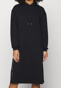 Nainen, joka on pukeutunut mustaan pitkähihalaiseen hupparimekkoon, jossa on sivuaukot ja valkoinen Calvin Klein Jeans -logo teksti sivuilla.