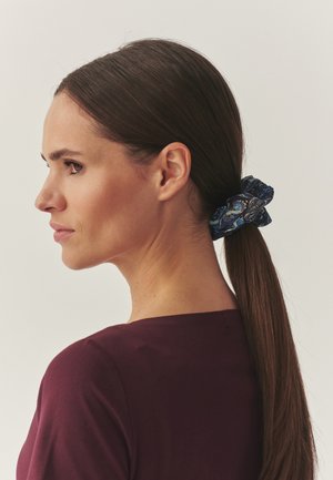 Coletero de satén en azul marino con estampados florales, diseño fruncido y textura suave, llevado en una coleta sobre cabello castaño oscuro.