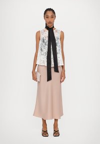White lace top features a high neckline with a black ribbon tie, paired with a long, blush skirt and black strappy shoes, holding a clutch.